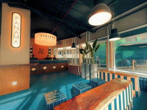 Un vestíbulo de hotel moderno con una mesa de recepción, un gran letrero iluminado que dice 'Panama Viajeros Hostels' y un amplio ventanal con vista al interior.