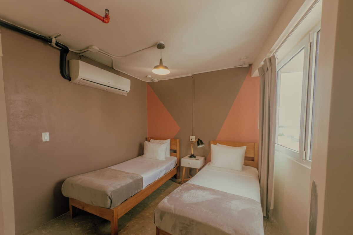 Una habitación con dos camas, una mesita de noche, una lámpara, un equipo de aire acondicionado y una ventana.