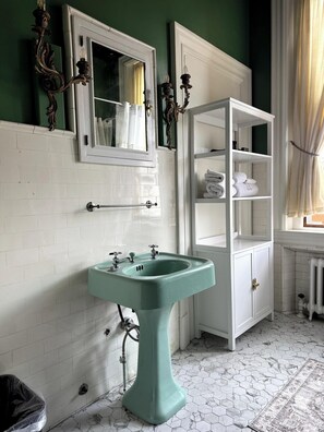 A bathroom with a white tiled wall, a green pedestal sink, a white cabinet with towels, and a window with curtains. - Villagio Luxury Shops And Hotel (Pittsburgh)