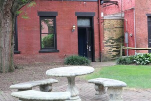 A red brick building with a black door, a window, and a small garden area with a stone table and benches.