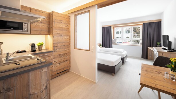 Una habitación de hotel moderna con kitchenette, una cama, un escritorio y un ventanal con cortinas.