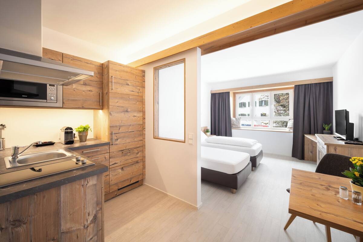 Ein modernes Hotelzimmer mit Küchenzeile, einem Bett, einem Schreibtisch und einem Fenster mit Vorhängen.