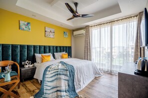 Une chambre d’hôtel comprenant un lit, une table de chevet, une chaise, une table, un ventilateur de plafond et une grande fenêtre avec des rideaux.