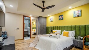 A bedroom with a bed, bedside table, and a desk with a chair. There is a mirror, a ceiling fan, and a door leading to a bathroom. - Lime Tree Hotel - Near Medanta Hospital (Gurugram)