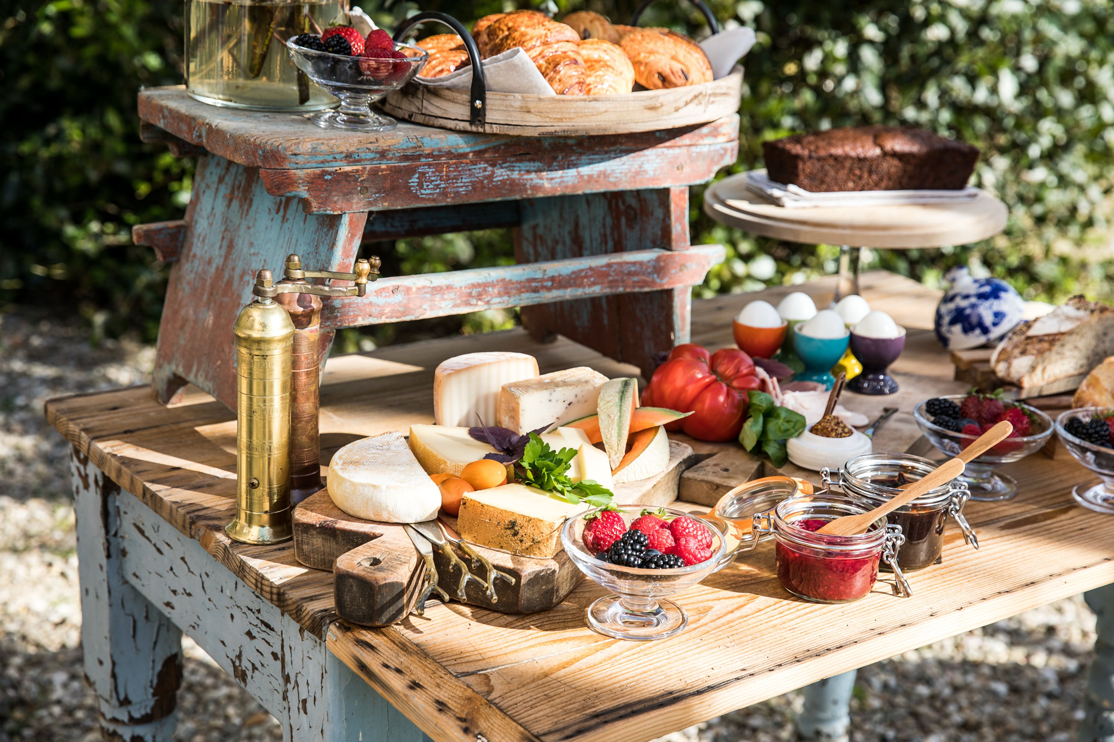 A rustic outdoor table with various cheeses, fruits, and pastries.