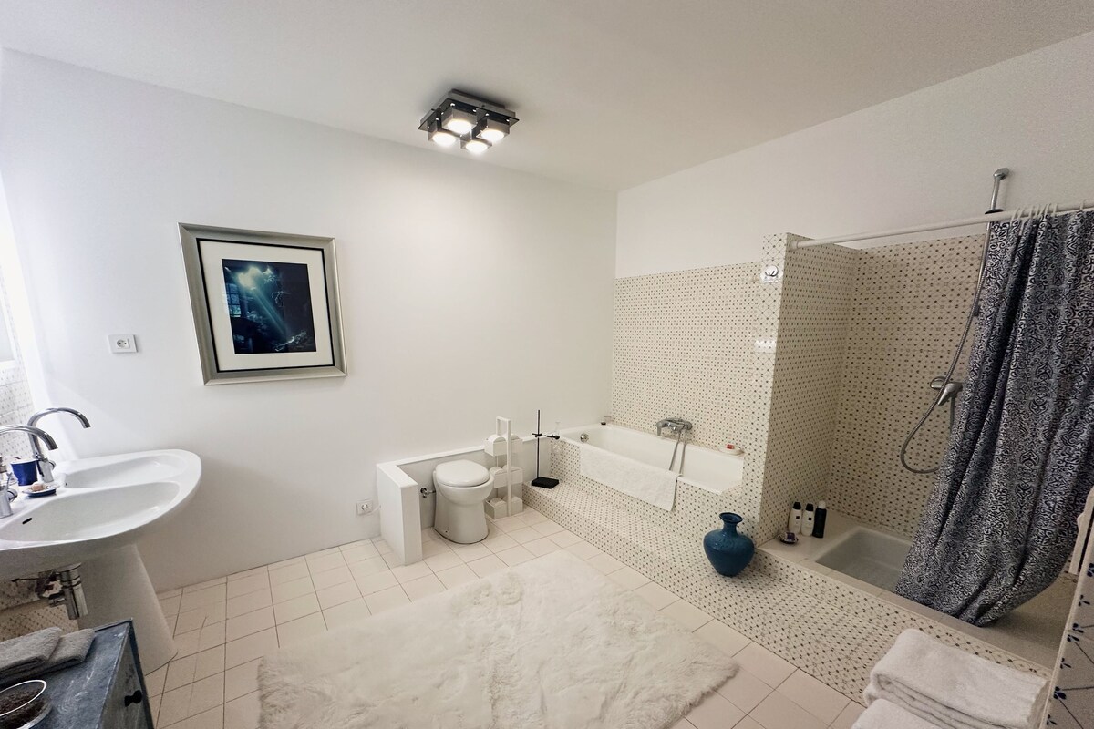 A modern bathroom with a toilet, sink, and bathtub. There is a framed picture on the wall, a white rug on the floor, and a shower curtain with a pattern.