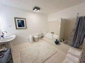 A modern bathroom with a toilet, sink, and bathtub. There is a framed picture on the wall, a white rug on the floor, and a shower curtain with a pattern. - Les Manibelles (Amarens)
