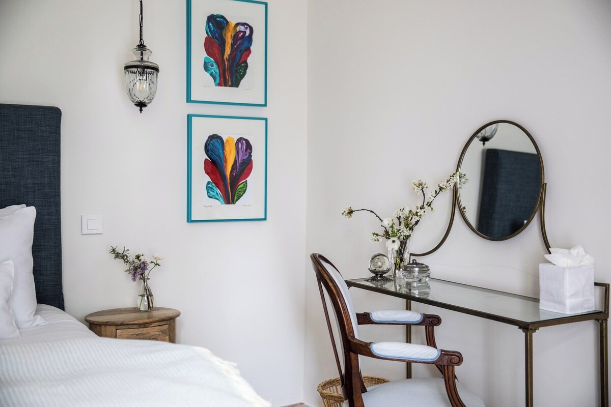 A hotel room with a bed, a bedside table with a vase of flowers, a chair, a desk with a mirror, and two framed artworks on the wall.