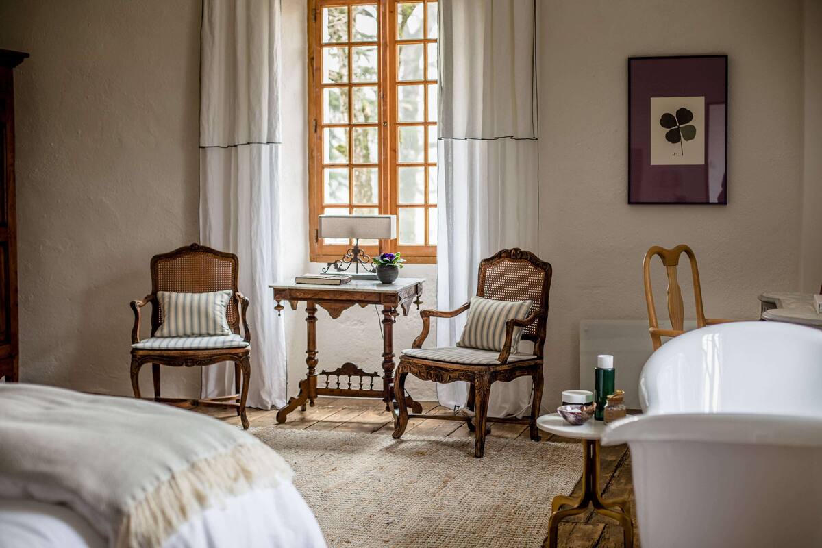 A room with two wooden chairs, a small table with a lamp, a window with curtains, and a framed picture on the wall.