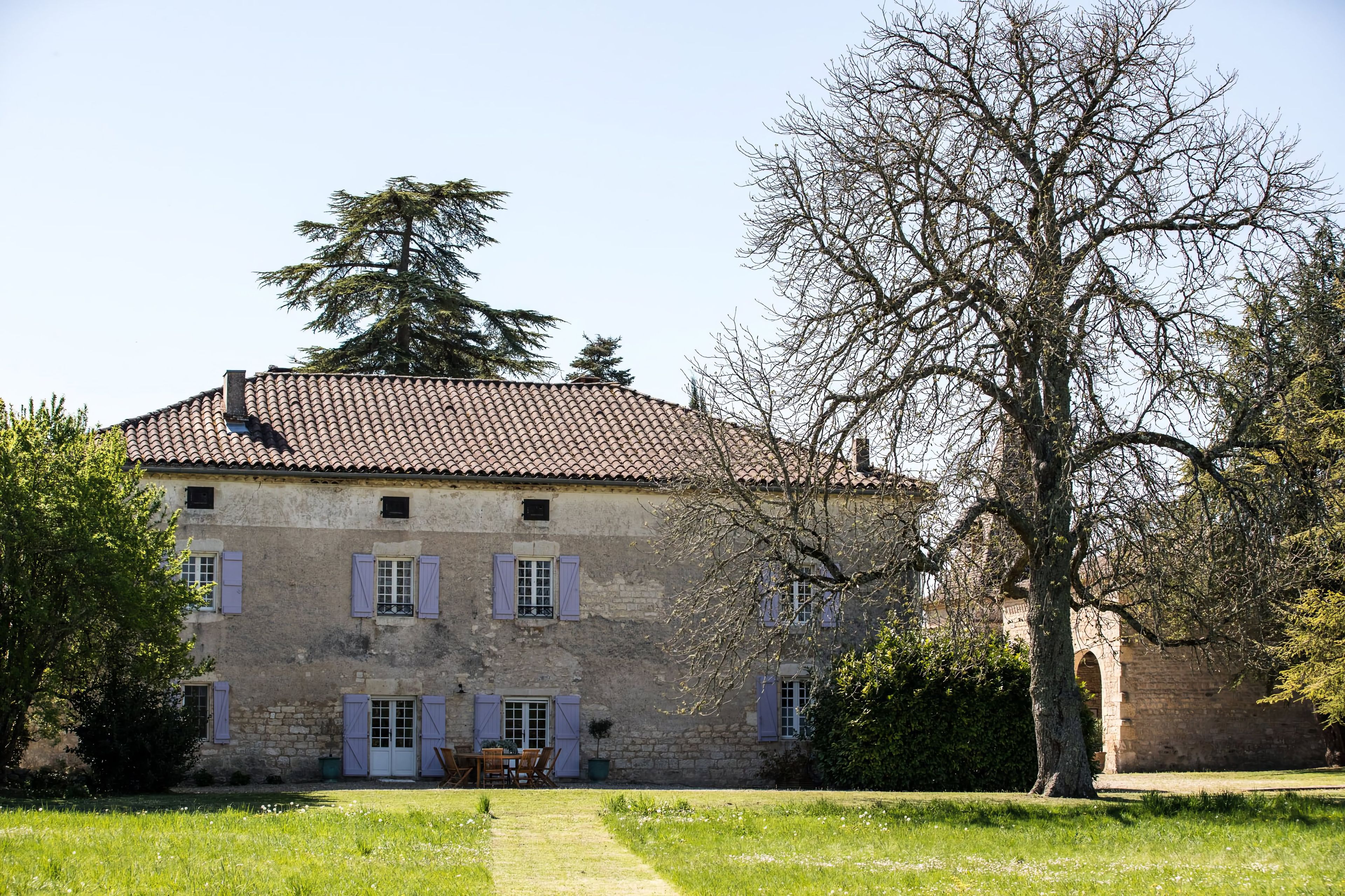 A stone building with a tiled roof, multiple windows with shutters, and a large tree in the foreground.