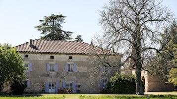 A stone building with a tiled roof, multiple windows with shutters, and a large tree in the foreground.