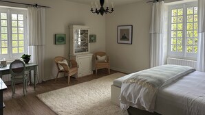 A bedroom with a bed, two wicker chairs, a white cabinet, a chandelier, and large windows with curtains. - Les Manibelles (Amarens)