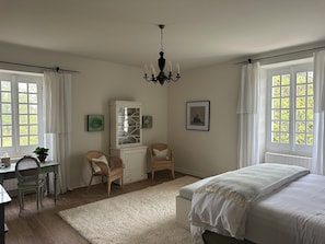 Ein Schlafzimmer mit einem Bett, zwei Korbstühlen, einem weißen Schrank, einer Chandelier und großen Fenstern mit Vorhängen.