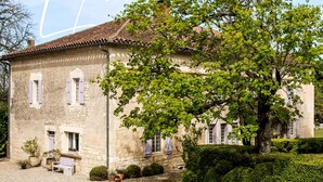 A stone building with a tiled roof, a large tree in front, and a gravel courtyard. - Les Manibelles (Amarens)