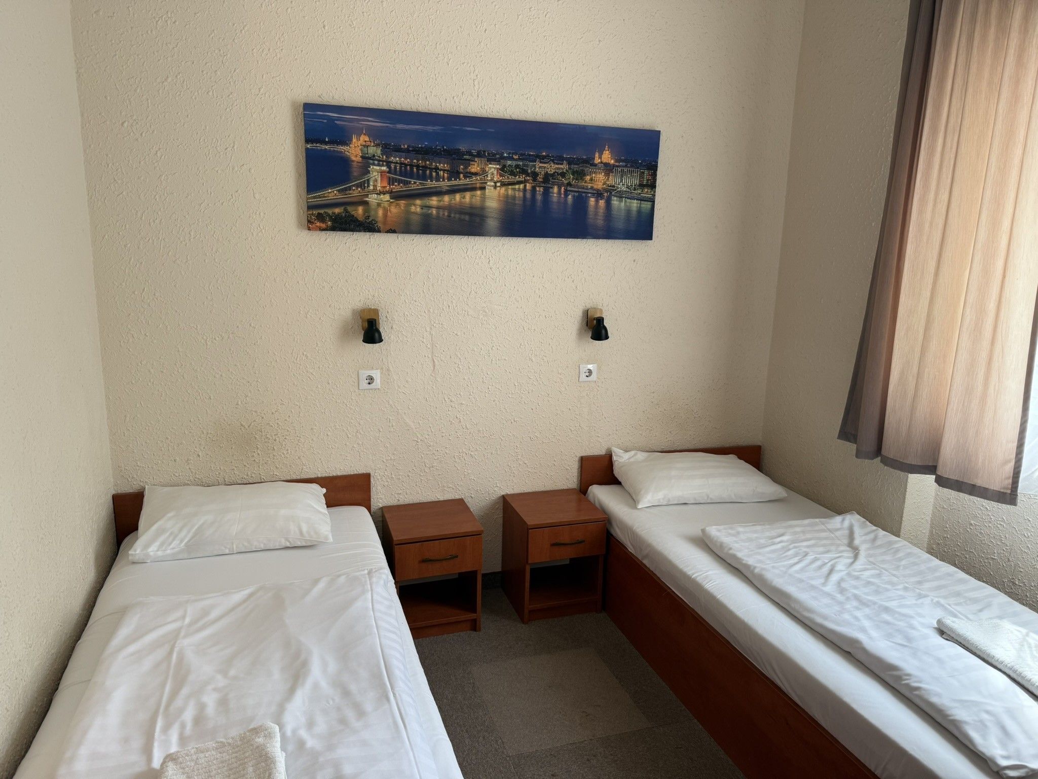 Habitación de hotel con dos camas individuales, una mesita de noche, una lámpara de pared y un cuadro de un paisaje urbano.