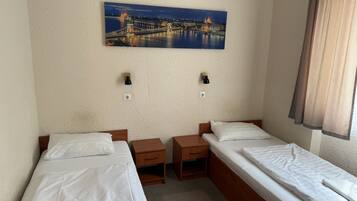 Habitación de hotel con dos camas individuales, una mesita de noche, una lámpara de pared y un cuadro de un paisaje urbano.
