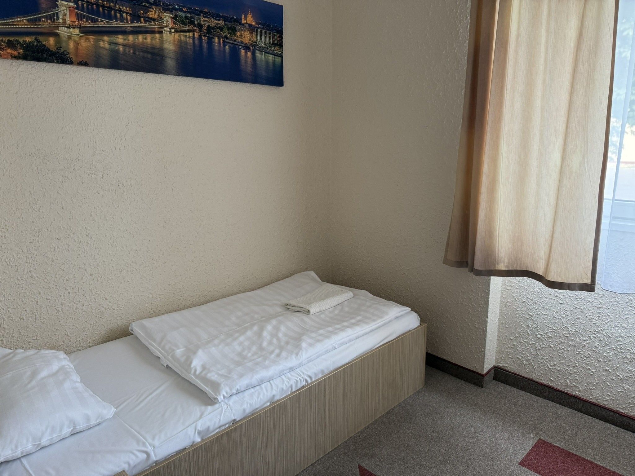 Una habitación pequeña con cama individual, una pared sencilla y un cuadro de un puente.