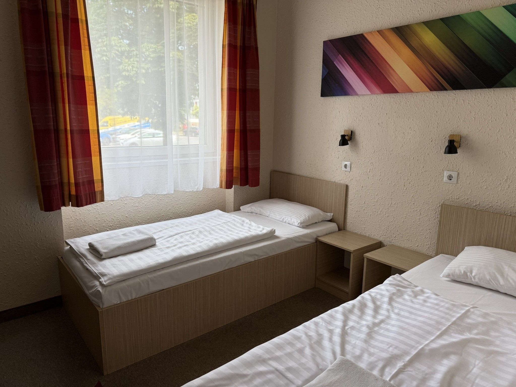 Habitación de hotel con dos camas, una ventana con cortinas translúcidas, una lámpara de pared y un cuadro.