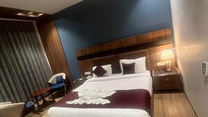 Une chambre d’hôtel moderne avec un grand lit, une tête de lit en bois, une table de chevet avec une lampe et un coin salon comprenant une chaise et une petite table.