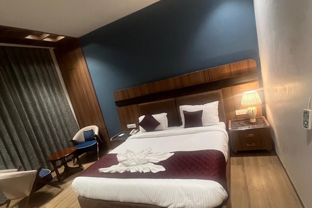 Une chambre d’hôtel moderne avec un grand lit, une tête de lit en bois, une table de chevet avec une lampe et un coin salon comprenant une chaise et une petite table.