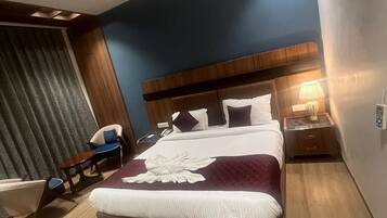 A modern hotel room with a large bed, a wooden headboard, a nightstand with a lamp, and a seating area with a chair and a small table.