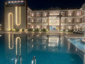 Un hôtel doté d’une grande piscine, un bâtiment moderne agrémenté de bandes lumineuses verticales, et une structure traditionnelle à plusieurs étages avec des balcons.