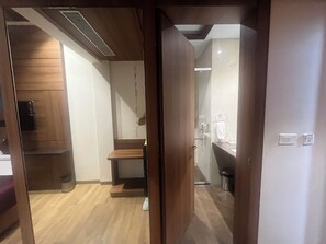 A hotel room with a wooden floor, a small wooden bench, a glass shower enclosure, and a sink with a towel hanging on the wall. - Nirvana Grand Hotel And Resort (Gaya)