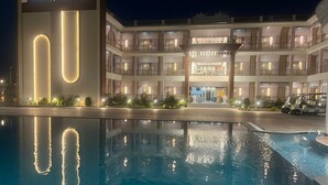 Un hôtel doté d’une grande piscine, un bâtiment moderne agrémenté de bandes lumineuses verticales, et une structure traditionnelle à plusieurs étages avec des balcons.