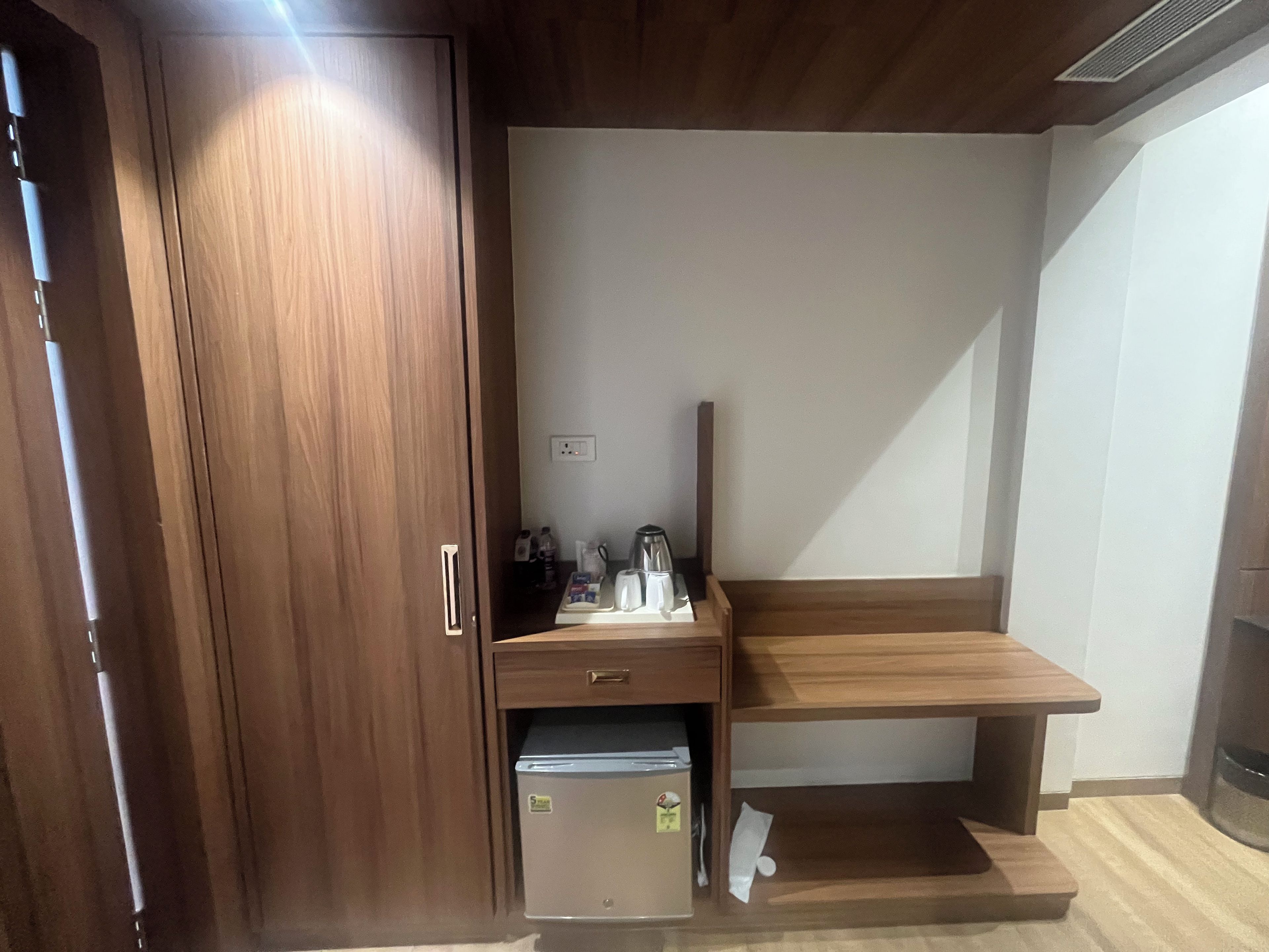 A compact room with a wooden wardrobe, a small desk with a chair, and a compact refrigerator.