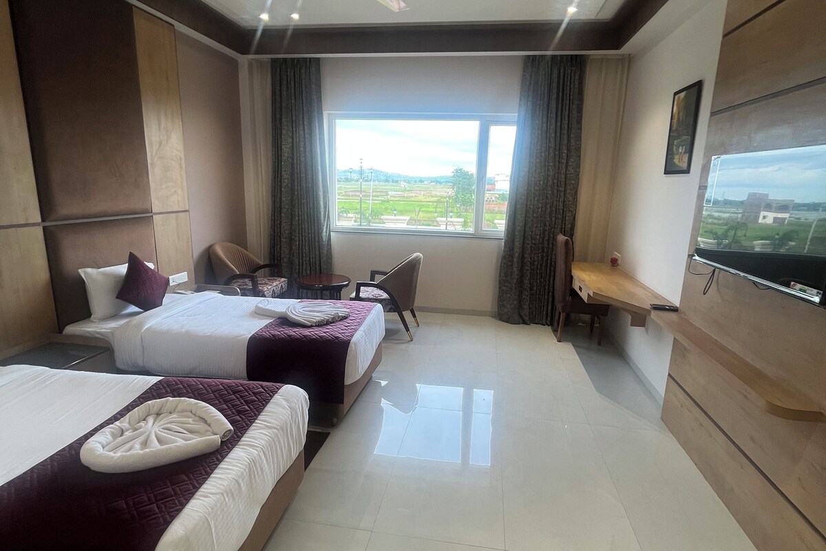 A hotel room with two beds, a desk, a chair, a TV, and a window with a view of the countryside.