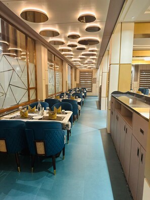 A long dining area with tables set for multiple guests, featuring a modern design with blue upholstered chairs and a central island with a counter. - Nirvana Grand Hotel And Resort (Gaya)