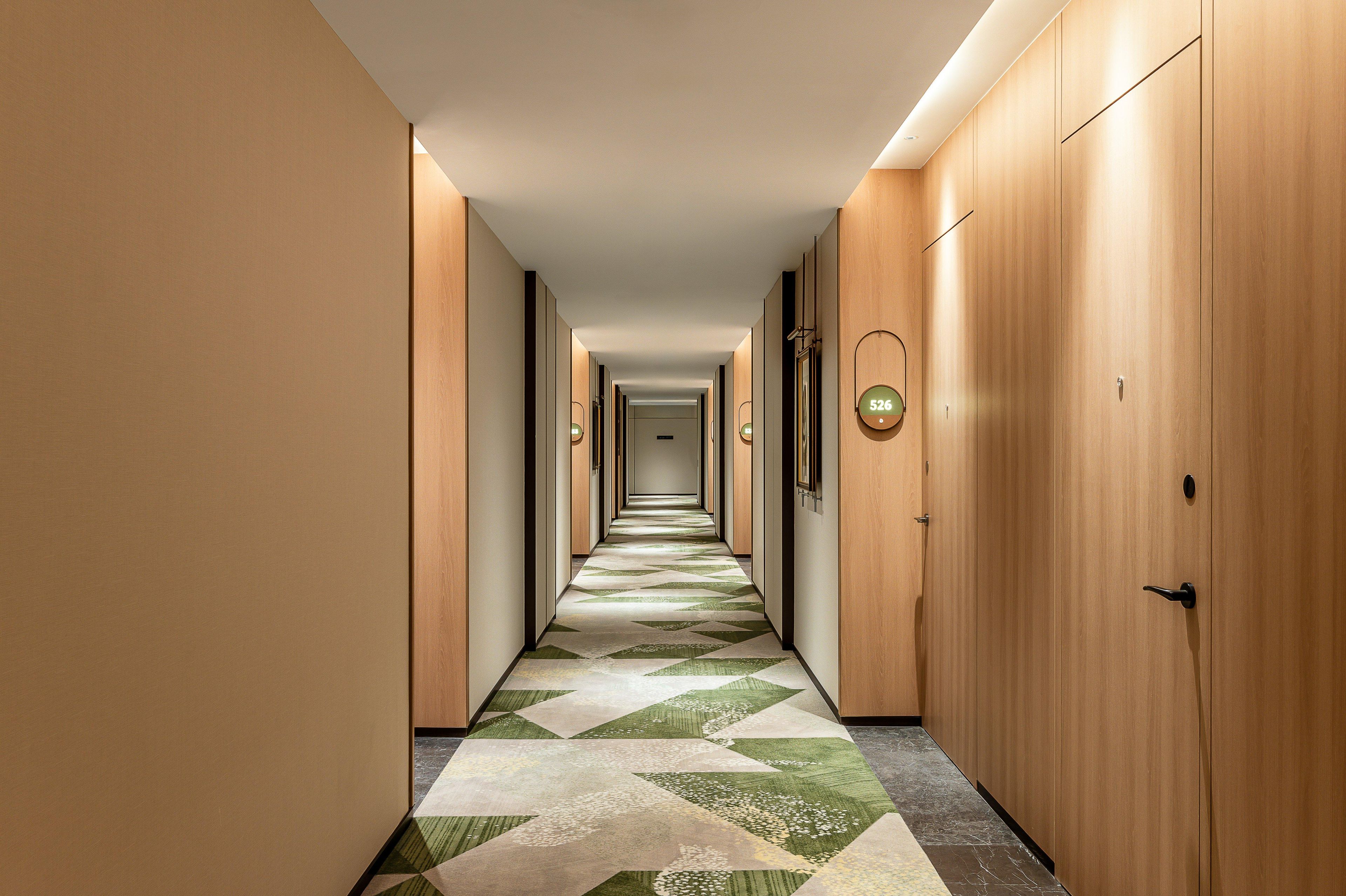 Un long couloir étroit d’hôtel, avec des portes en bois, un sol à motifs et un luminaire au plafond.