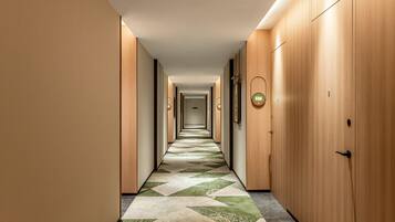 Un long couloir étroit d’hôtel, avec des portes en bois, un sol à motifs et un luminaire au plafond.