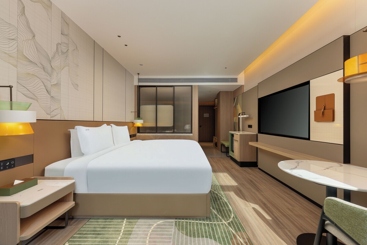 A modern hotel room with a large bed, bedside tables, a desk, a chair, a TV, and a small table.