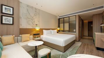 A modern hotel room with a large bed, a sofa, a small table, and a green ottoman. There are framed pictures on the wall and a large window with blinds.