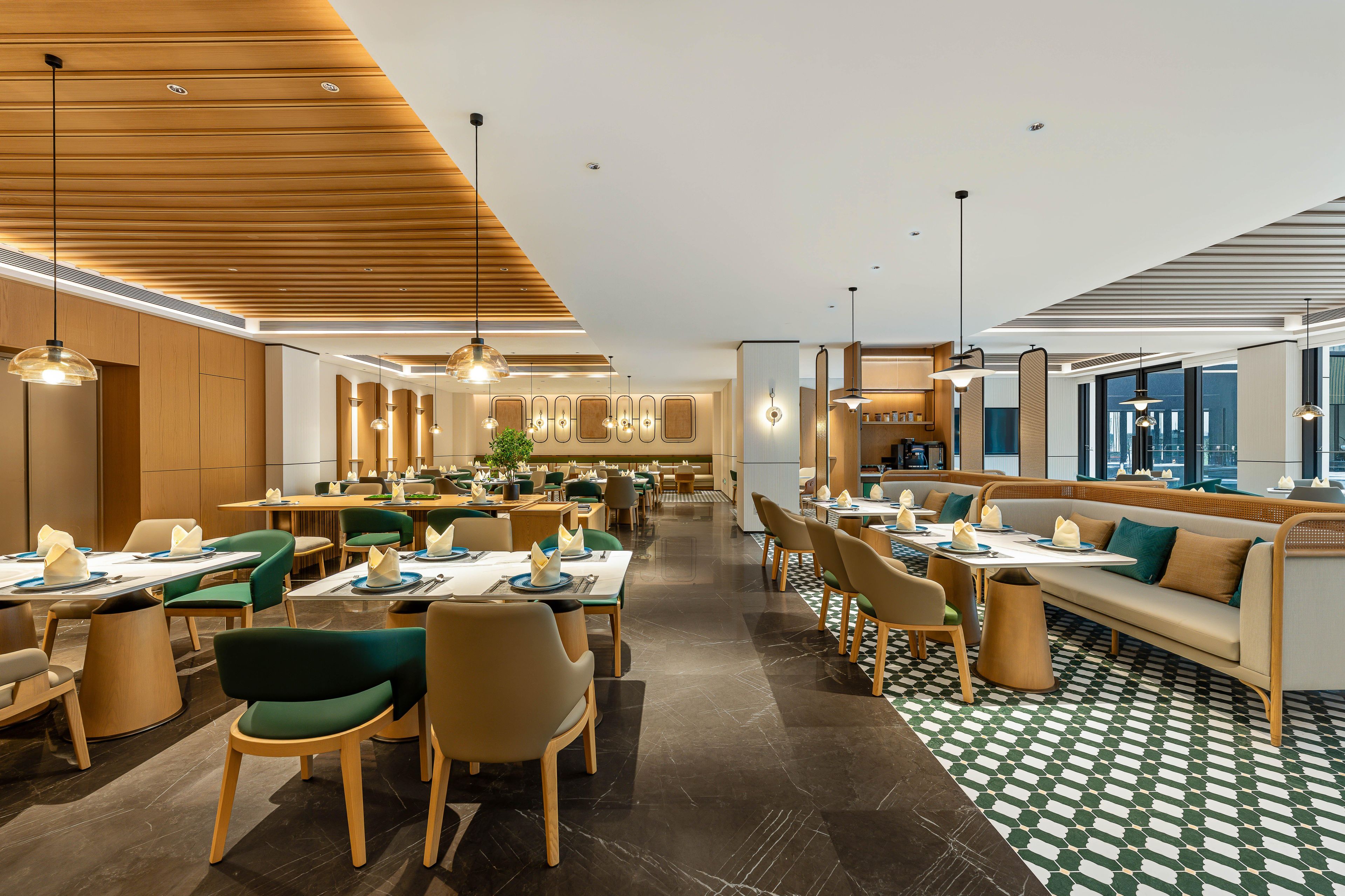 A modern hotel dining area with wooden ceiling panels, green and beige chairs, and a checkered floor pattern.