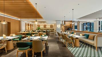 A modern hotel dining area with wooden ceiling panels, green and beige chairs, and a checkered floor pattern.
