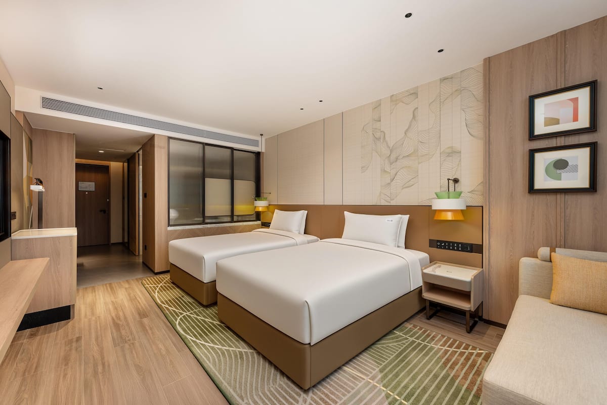 Ein Hotelzimmer mit zwei Betten, einem Schreibtisch, einem Stuhl, einer Lampe und einer Wand mit Kunstwerken.
