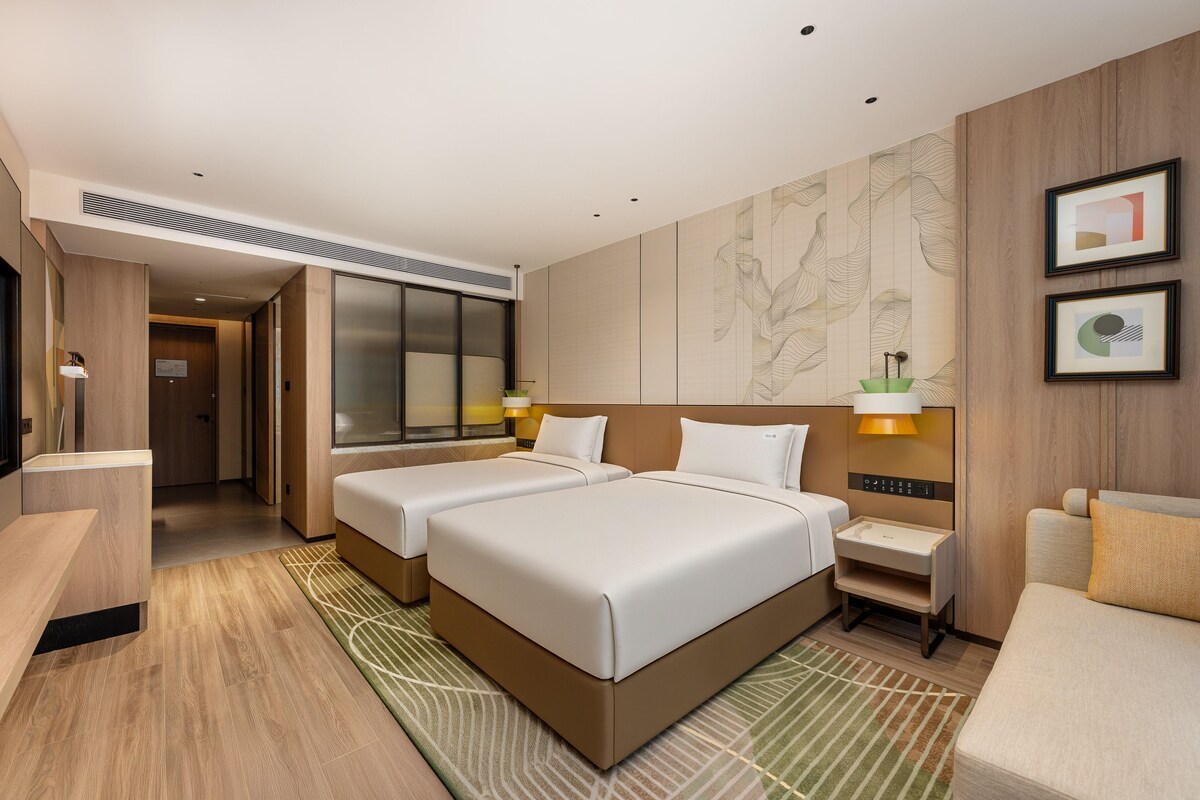 A hotel room with two beds, a desk, a chair, a lamp, and a wall with artwork.