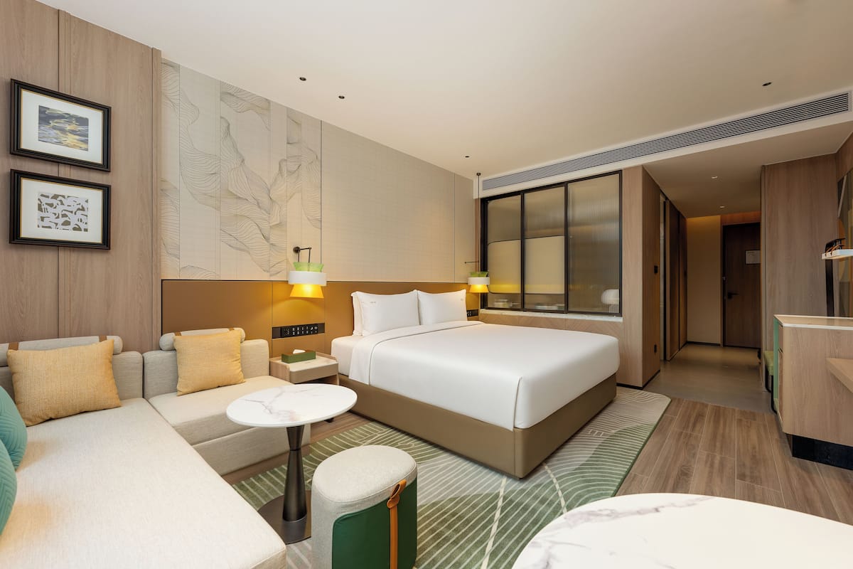 Ein modernes Hotelzimmer mit einem großen Bett, einer Couch, einem kleinen Tisch und einem grünen Pouf. An der Wand befinden sich gerahmte Bilder und ein großes Fenster mit Jalousien.