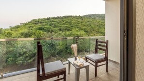 A balcony with two wooden chairs and a small table, overlooking a lush green hillside. - Alaya Stays By Elivaas Lake City Heights (Udaipur)