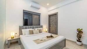 A modern bedroom with a large bed, bedside tables with lamps, a potted plant, and a window with a view. - Alaya Stays By Elivaas Lake City Heights (Udaipur)