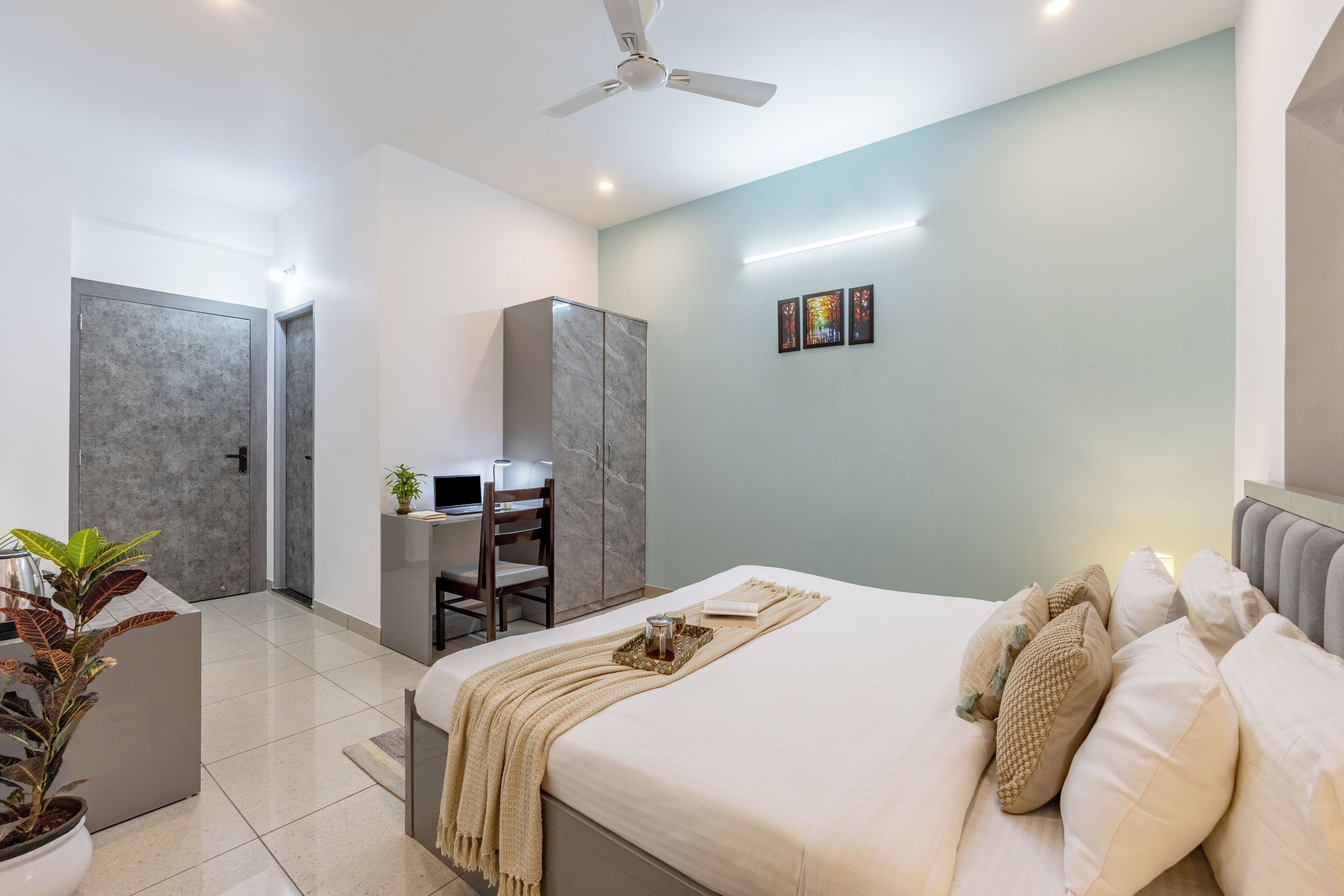 Une chambre à coucher moderne avec un lit, une table de chevet et un bureau avec une chaise. On y trouve également un ventilateur de plafond, une plante en pot et une étagère fixée au mur.