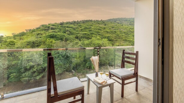 A balcony with two wooden chairs and a small table, overlooking a lush green landscape. - Alaya Stays By Elivaas Lake City Heights (Udaipur)