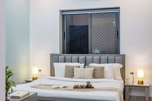 A modern bedroom with a large bed, two bedside tables, a window with a view, and a tray with tea and sugar. - Alaya Stays By Elivaas Lake City Heights (Udaipur)