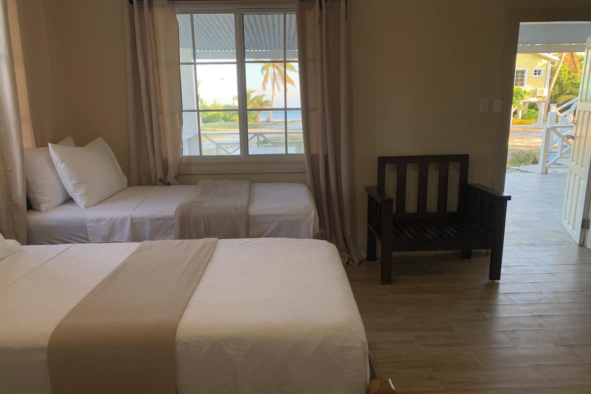 A hotel room with two single beds, a wooden bench, and a view of the beach and palm trees through the window.