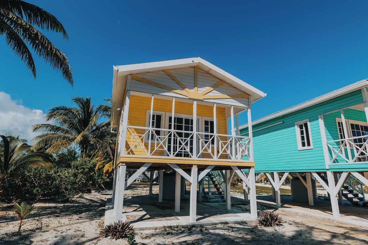 A two-story beach house with a balcony, surrounded by palm trees and a clear blue sky.