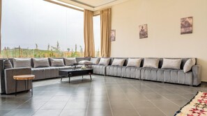 A modern living room with a grey sectional sofa, a small round table, and a coffee table. There are large windows with curtains, a tiled floor, and three framed artworks on the wall. - Life Resort (Marrakech)
