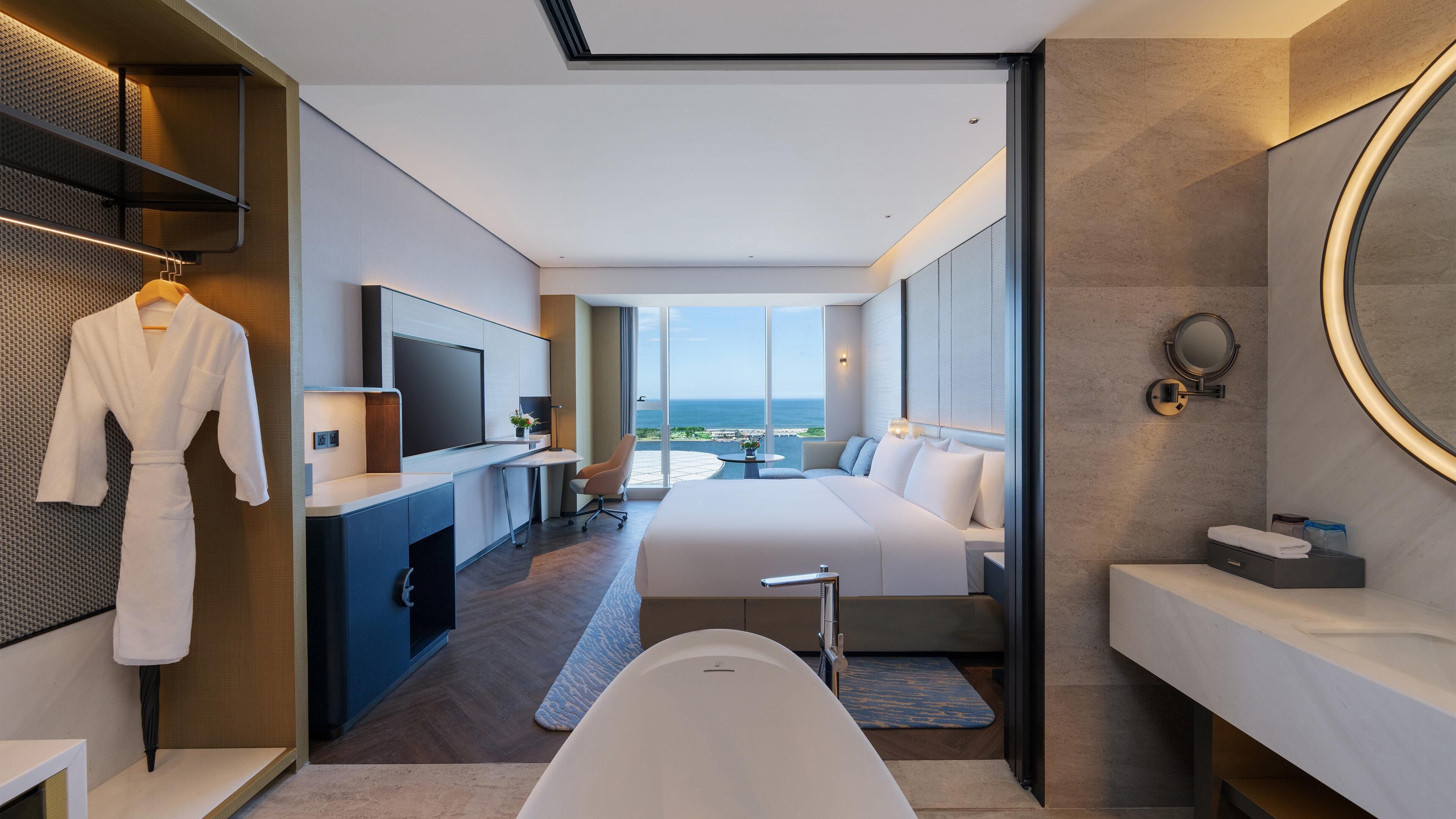 Una habitación de hotel moderna con una cama grande, una zona de estar, un televisor, una bañera y vistas al océano.
