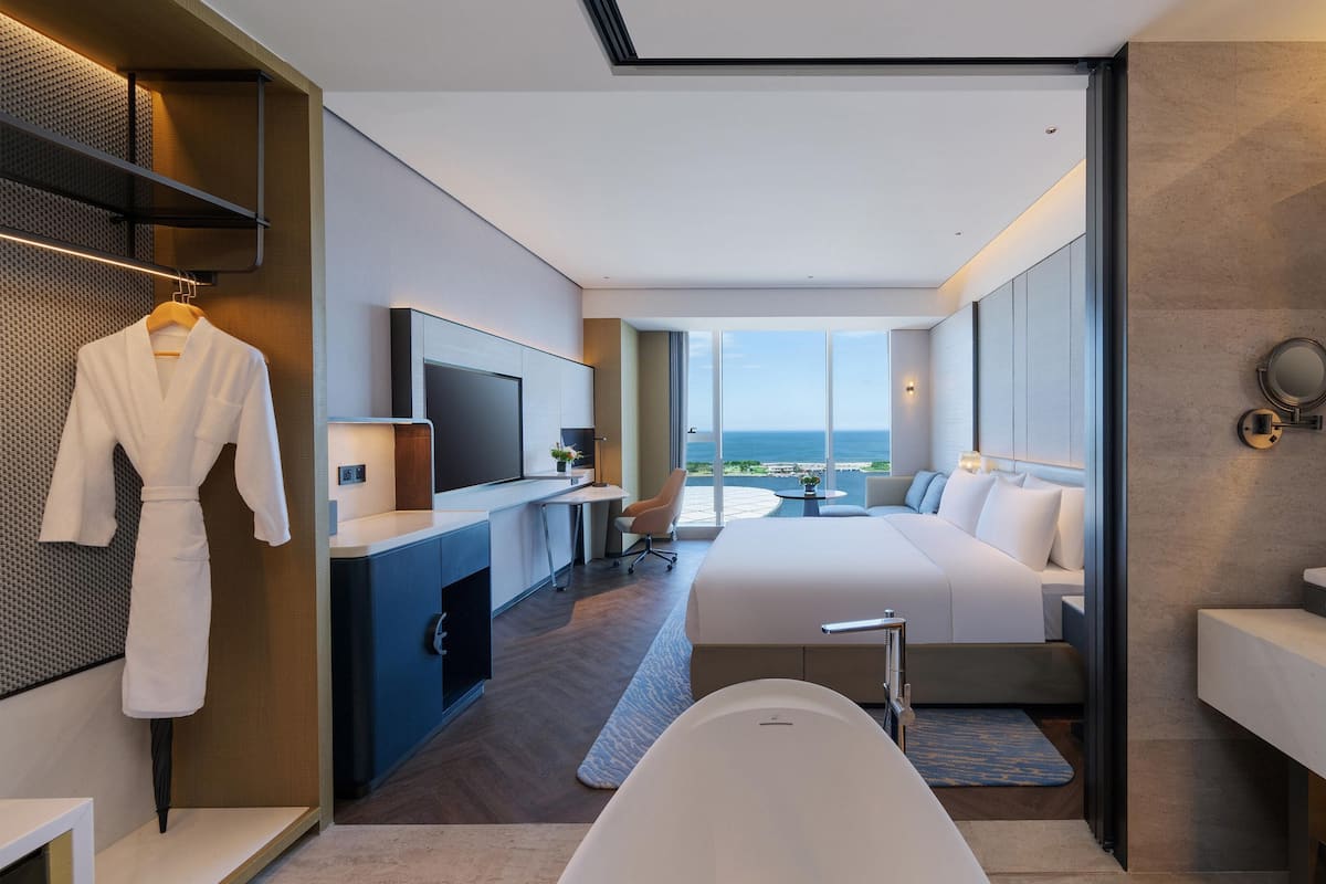Une chambre d’hôtel moderne dotée d’un grand lit, d’un coin salon, d’une télévision, d’une baignoire et offrant une vue sur l’océan.